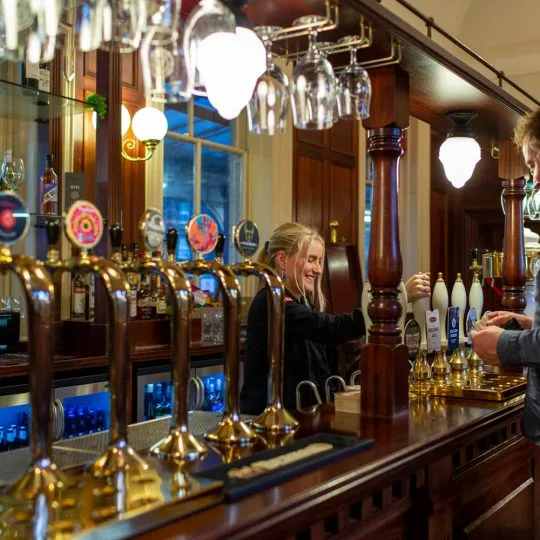Barmaid pulling a pint behind a row of brass and wood handpulls