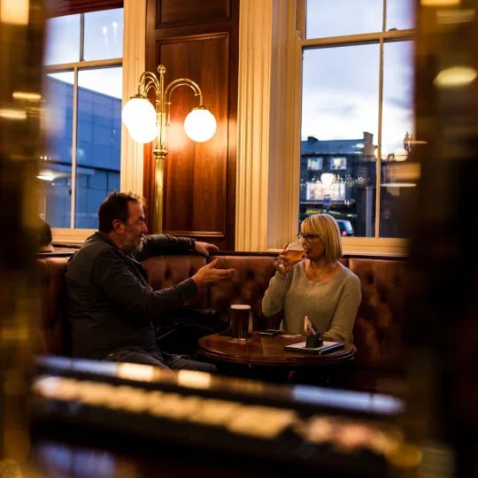 Two drinkers chatting in a leather booth by the window at Harrogate Tap